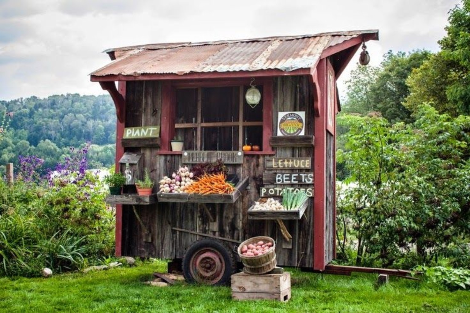 Rustic wooden farm stand with red trim and fresh vegetables beside a garden